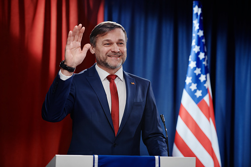 Waist up portrait of mature man giving oath while standing at podium on stage against red curtain, copy space