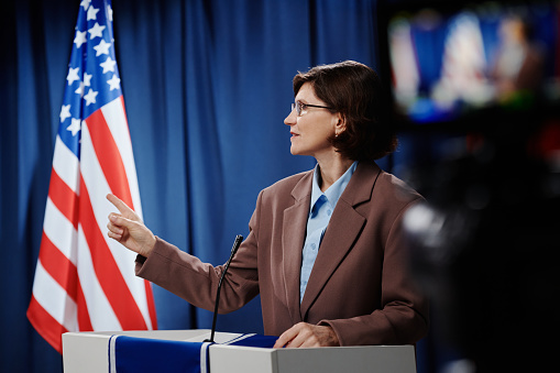 Mature female political leader looking at opponent and pointing at him while standing by platform and expressing her opinion