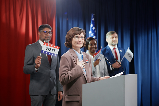 Mature smiling woman keeping hand on chest and looking at electorate while standing by platform and speaking in microphone