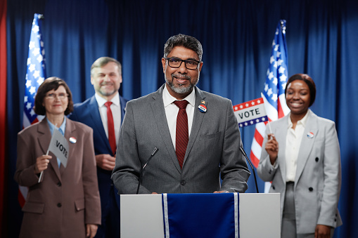 Mature multiethnic male candidate making speech during voting campaign and looking at camera while standing in front of public