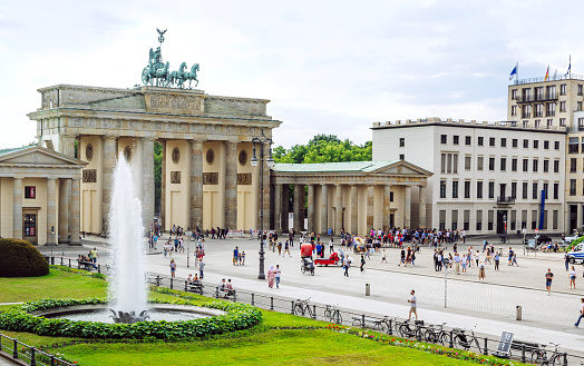 A vibrant scene at Brandenburg Gate in Berlin, Germany, bustling with tourists and locals. The iconic landmark stands prominently, surrounded by people, greenery, and a beautiful fountain.