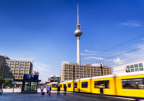 This striking image captures a bustling Berlin scene featuring the famous TV Tower, a yellow tram in motion, and modern urban architecture under a clear blue sky.