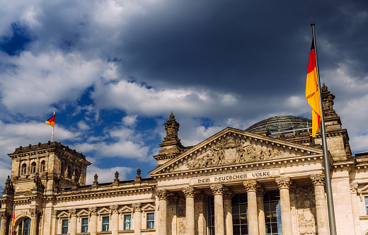 Striking image of the Reichstag building in Berlin under a dramatic cloudy sky. The German flag flutters proudly, encapsulating a sense of national pride and history.