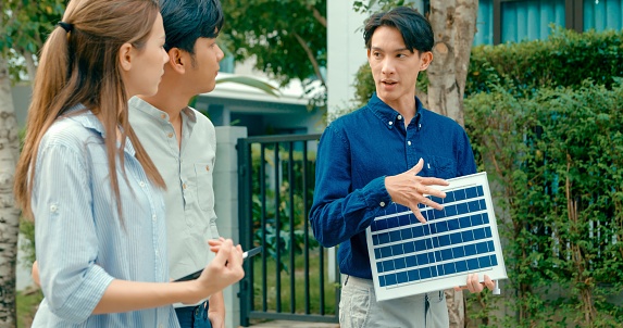 Asian real estate agent explains solar panel benefits to a potential buyer outside a residential home. Consulting Eco-Friendly Home with Real Estate Agent Concept.