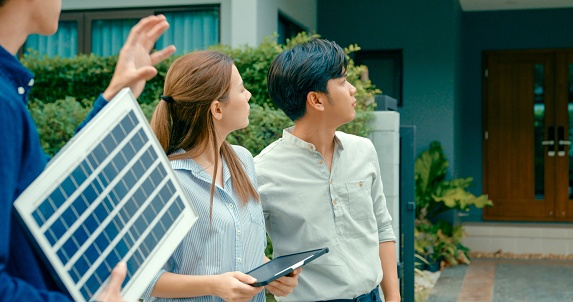 Asian real estate agent demonstrates solar panel benefits to interested young couple outside a modern home. Consulting Eco-Friendly Home with Real Estate Agent Concept.