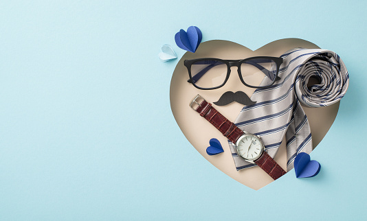 Fathers' Day greeting card concept. Top view layout of classy necktie, wristwatch, glasses, mustaches, and paper hearts, in heart-shaped frame on pastel blue with empty space for message
