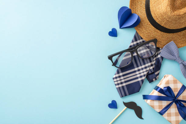 Pay tribute to Dad with delightful top view composition featuring straw hat, refined necktie, ribbon-wrapped gift box, glasses, mustache, hearts set on pastel blue backdrop, great for greeting message