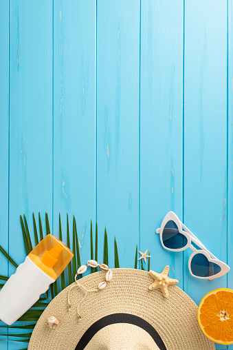 A vertical vibrant display of summer vacation essentials, including a straw hat, sunglasses, sunscreen, and fresh orange, arranged on a blue wooden surface, symbolizing travel and leisure