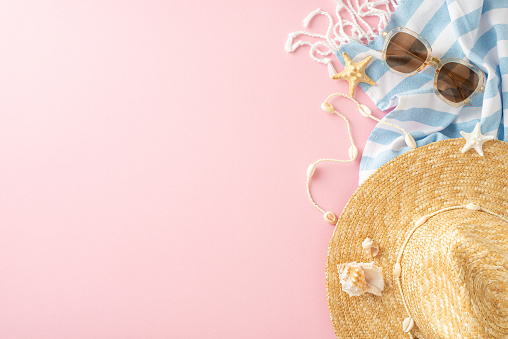 A top view of summer vacation accessories, featuring a straw hat, sunglasses, and a blue striped towel adorned with starfish on a pink background