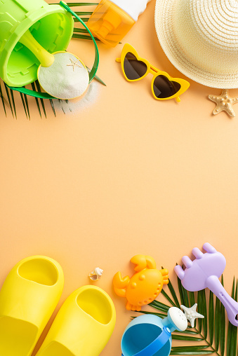 Lively holiday vertical top view scene: kids' sand playset, SPF protection, chic sunhat, shades, cute slippers, palm leaves and more, arranged on soft orange background, ideal for advertising material