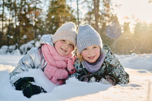Pretty little girls in winterwear looking at camera with smiles on sunny winter day in natural environment while having fun in snowdrift