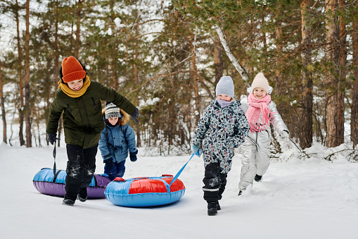 Group of cute cheerful children in winterwear running down forest road on winter day and laughing while pulling their snow tubes