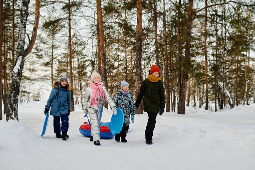 Four cheerful intercultural kids in warm winter jackets and beanie hats pulling snow tubes and carrying slides while walking along road