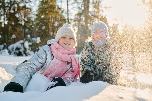 Two cheerful girls in winterwear lying in snowdrift and looking at camera while throwing snow and enjoying sunny winter day in park
