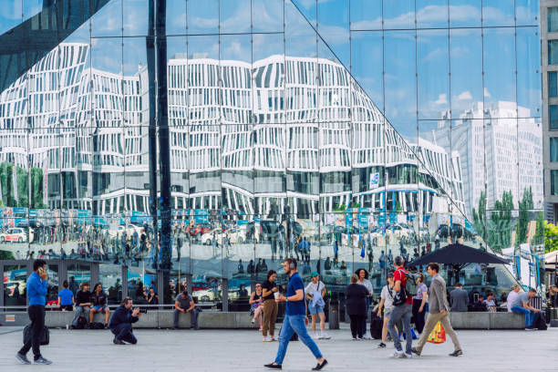 Berlin, Germany - June 07, 2023: Diverse crowd of people walking by a highly reflective building in Berlin, Germany, showcasing urban life and modern architecture.