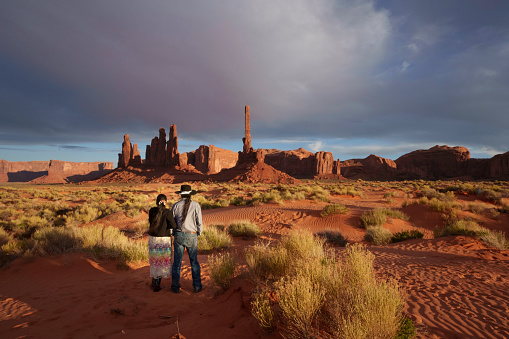 Traditional Navajo Married Couple Viewing Totem Pole Rock in Monument Valley Tribal Park at Dusk with Cloudscape and Patterns in the Sand
