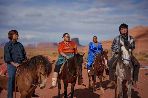 Young Navajo Family Riding Horseback on Their Ranch in Monument Valley Arizona at Dusk with Majestic Red Rock Mountains East and West Mittens and Merrick Butte in Background and Dramatic Cloudscape