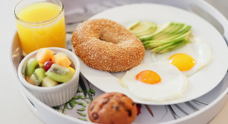 Breakfast, orange juice and food in plate closeup for nutrition or wellness with cupcake on tray in home in the morning. Eggs, bagel and drink for healthy diet with fruits, avocado slice or muffin