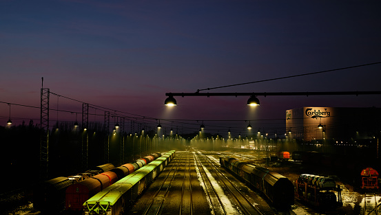 Fredericia, Denmark. January 12, 2024. High angle view of lights illuminating over trains parked on railway tracks against blue sky at night