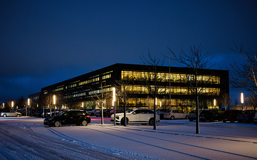 Fredericia, Denmark. January 17, 2024. Cars parked on snow covered parking lot in front of modern Illuminated office building against cloudy blue sky at night