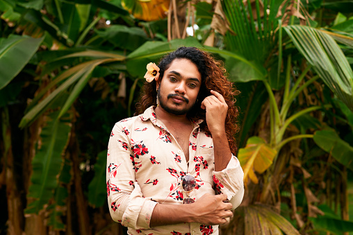 Reflective of LGBTQ pride, gender fluidity, self-expression. Sri Lankan man with floral shirt, hair flower poses in garden. Gentle gaze, vibrant background. Celebrates diversity, beauty in nature.