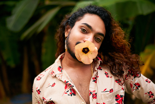 Person represents LGBTQ pride, cultural diversity, gender fluidity vibrant foliage. South Asian man shirt, expressive eyes holds yellow flower in mouth, bold femininity, self-love in rich garden.