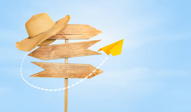wooden road sign with a beach hat on the edge and a flying paper airplane against the sky. travel concept
