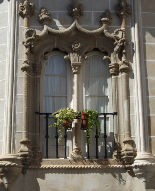 This majestic balcony in Evora Portugal is framed by two lateral columns and a central column, forming a double arch for a pair of glass doors that open onto a railing. The top is adorned with stone figures, enhancing its charm.