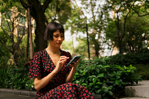 Young asian woman using mobile phone in nature. Female in casuals using smart phone sitting at park.