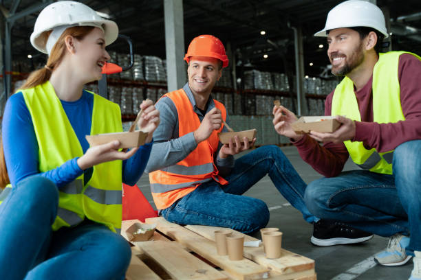 istock Group smiling workers wearing hard hats, vests, holding lunch boxes, eating lunch, sitting, talking 2149876956