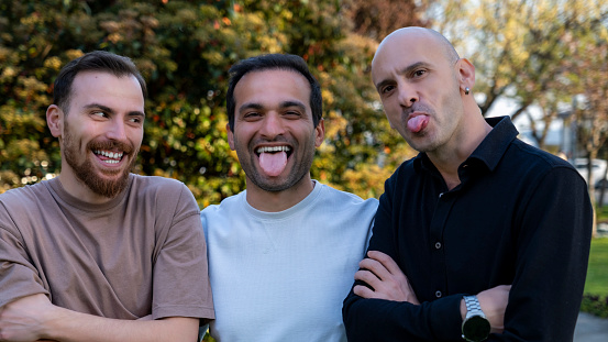 In this close-up portrait captured in a wooded area, three middle-aged men are seen goofing around and making silly faces, enjoying a moment of lightheartedness. Their playful expressions add a touch of humor to the serene backdrop of the forest, showcasing the joy of camaraderie and friendship