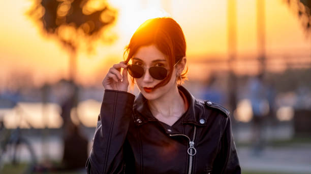 A beautiful woman sitting on a bench by the sea poses with sunglasses, her gaze focused on the endless horizon as the last rays of the sun illuminate the sky. With the shimmering seascape behind, this moment blends seamlessly with the serenity of the coast and the beauty of nature.