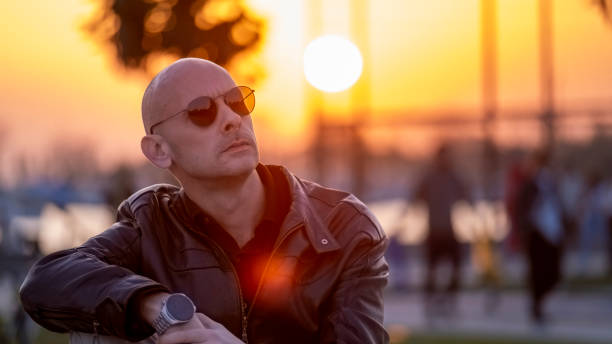 Sitting on a bench by the sea, a charismatic man strikes a pose with sunglasses, his gaze fixed on the infinite horizon as the last rays of the sun illuminate the sky. With the shimmering sea view behind him, this moment blends seamlessly with the tranquility of the shore and the beauty of nature
