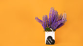 Bouquet of fresh lupine flowers in white vase shaped like bag and hello summer lettering with smile on black heart on orange background