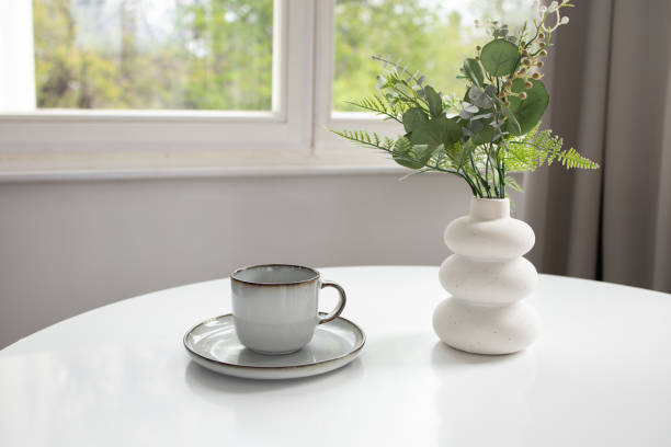 istock White vase with artificial greenery and empty cup on white table and window at the back 2149892127