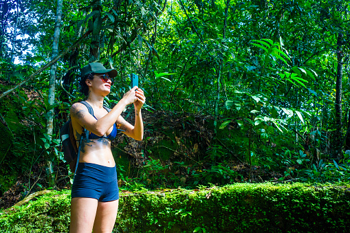 woman photographs nature with her cell phone. wears bathing suit, cap, sunglasses and backpack