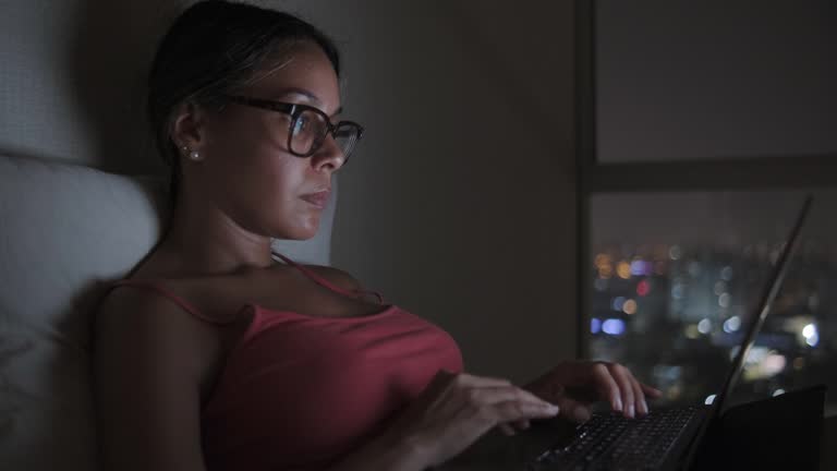 A woman is typing on a laptop in a dimly lit room, background view city lights in bokeh, Digital nomad concept.