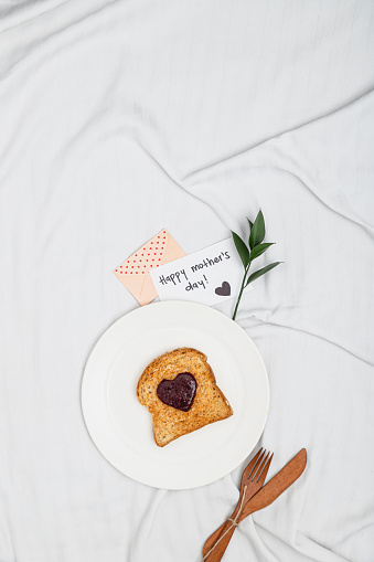 Happy mother's day greeting card on a wrikled sheet bed with a heart shaped jam on a toast over round plate with greeting card, cutlery and a little green twig with space for text and copy