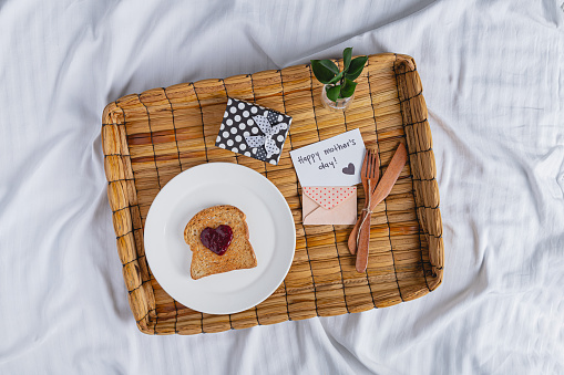 A rustic basket tray with happy mother's day greeting card, a little gift in a box, and a toast of bread with heart shape made out of jam, all over a bed on wrinkled sheet