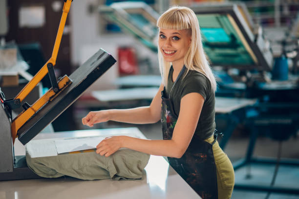 Portrait of smiling female print shop worker using heat press on tee. Portrait of a smiling female entrepreneur standing at printing workshop and using heating press on silkscreen printed t-shirt. Happy female graphic technician preparing t-shirt for heating press. t shirt transfer stock pictures, royalty-free photos & images