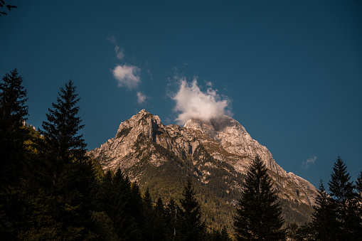 A serene mountain landscape featuring towering peaks embraced by wispy clouds and framed by coniferous trees under a clear azure sky.