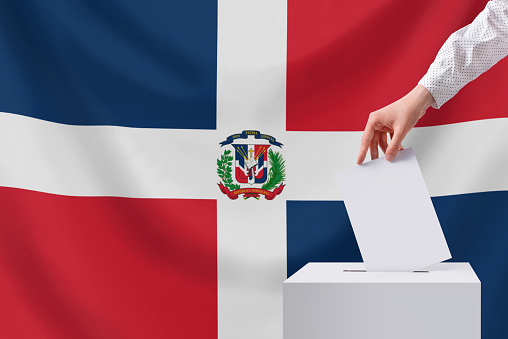 The voter throws the ballot into the box. Elections, Dominican Republic. Dominican Republic flag in the background.