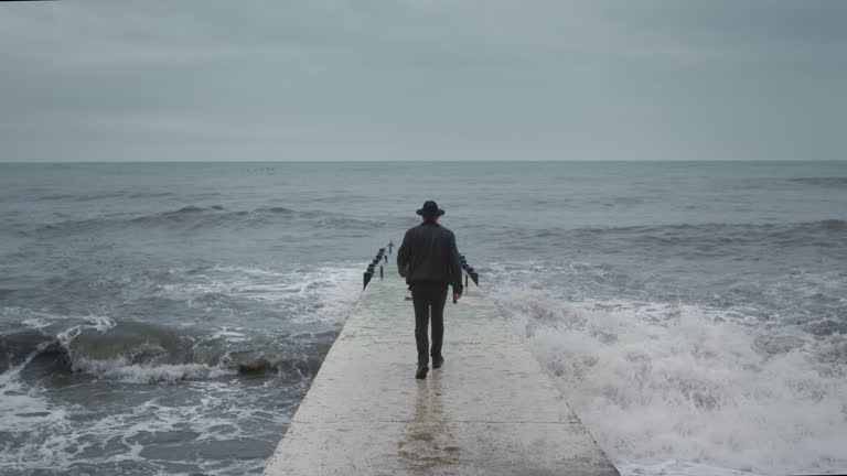 Man in a jacket and hat stand on a breakwater. Male on the seawall, big waves, artist in a rock band