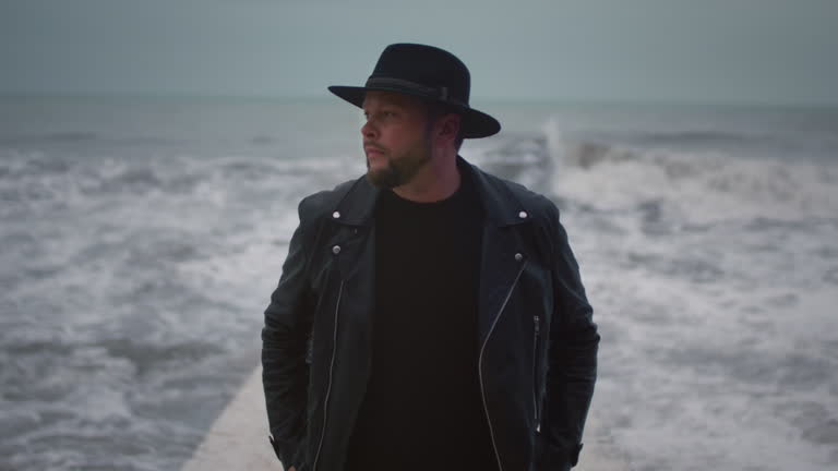 Man in a jacket and hat stand on a breakwater. Male on the seawall, big waves, artist in a rock band