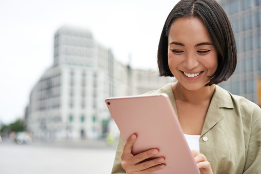 Retrato de mujer asiática leyendo, usando tableta mientras está de pie en la calle, sonriendo mientras mira la pantalla photo