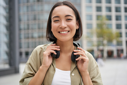 Retrato de una chica asiática sonriente con auriculares, posando en el centro de la ciudad, escuchando música photo