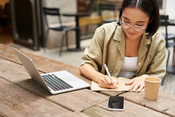 istock Portrait of concentrated girl doing homework, attending online course, making notes while working on laptop from cafe 2149904602