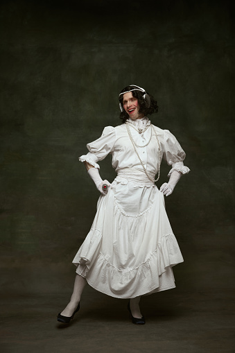 Full-length portrait of young man dressed as woman, medieval person and dancing in headphones against vintage studio background. Concept of self-expression, comparisons of eras, freedom, human rights.