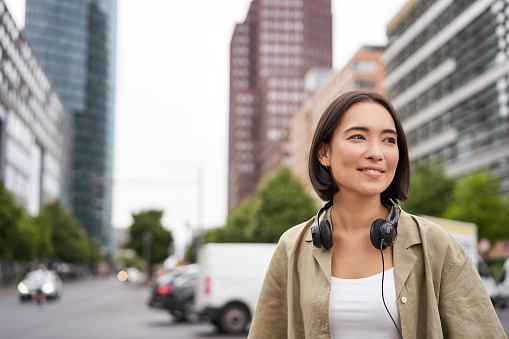 Retrato de una joven mujer asiática con auriculares, posando en la ciudad, sonriendo y mirando hacia otro lado, de pie en la calle del centro de la ciudad photo