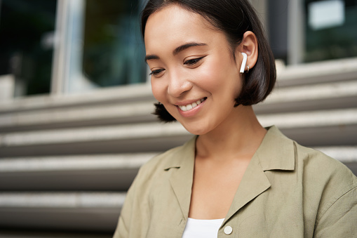 Retrato de una chica asiática sonriente que escucha música, podast en auriculares inalámbricos, usando auriculares al aire libre, sentada en la calle photo